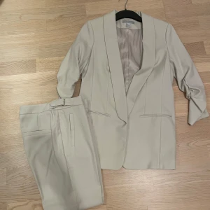  kostym med kavaj och byxa - Snygg beige kostym från H&M med kavaj och matchande byxa. Kavajen har 3/4-ärm med rynkdetalj och stilren krage, medan byxan har ett bälte med guldfärgat spänne. Materialet är mjukt och lätt, perfekt för dig som gillar en clean look. Använd 1 gång 