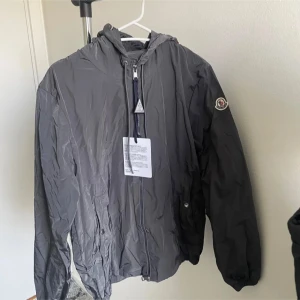 Grå Moncler vindjacka med huva - Säljer en grå vindjacka från Moncler med huva och dragkedja framtill. Jackan har Moncler-logga på ärmen och snygga röd, vit och blå ränder på huvan. Perfekt för blåsiga dagar och enkel att matcha med det mesta.