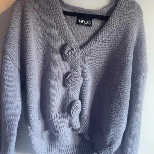 Ljuslila stickad kofta från Pieces - Supermysig ljuslila stickad kofta från Pieces med stora virkade knappar framtill och v-ringad hals. Koftan har en loose passform och långa ärmar med breda muddar. Perfekt att slänga på sig när det är lite kyligt ute. Nästan helt ny!