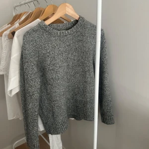 Grå stickad tröja med rund hals - Mysig grå stickad tröja med rund hals och långa ärmar. Tröjan har en melerad look och ribbade muddar vid ärmslut och nederkant. Perfekt för lager-på-lager under kyliga dagar.