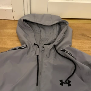 Grå vindjacka från Under Armour - Snygg grå vindjacka från Under Armour med svart logga på bröstet och svarta dragsnören i huvan. Jackan har hel dragkedja, ventilerande meshpaneler och elastiska muddar. Perfekt för dig som gillar sportig stil och vill ha något lätt och funktionellt.