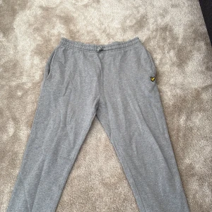 Grå mjukisbyxor från Lyle & Scott - Snygga grå mjukisbyxor från Lyle & Scott med elastisk midja och snörning. Byxorna har sidofickor, en bakficka och den klassiska gula loggan broderad på benet. Perfekta för chill eller träning, mjukt bomullsmaterial som är skönt mot huden.