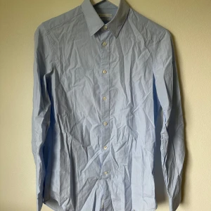 Ljusblå slim fit skjorta från Arket - Snygg ljusblå skjorta från Arket i slim fit-modell. Tillverkad i tunn och mjuk bomullspoplin, med klassisk krage och vita knappar framtill. Långärmad och stilren design som passar perfekt till jeans eller chinos. 1F3