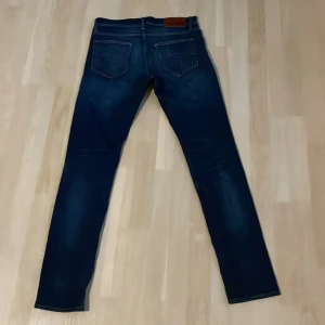 Mörkblå jeans från Tiger of Sweden - Snygga mörkblå jeans från Tiger of Sweden med klassisk femficksdesign och diskreta slitningar. Jeansen har en slim passform och raka ben, perfekt för en stilren look. Tillverkade i mjukt jeansmaterial med snygga kontrastsömmar.