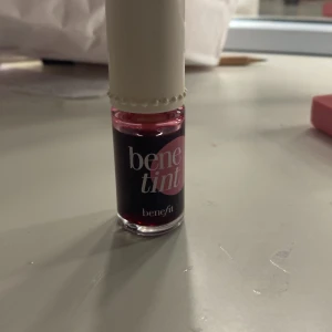 Benefit Benetint lip & cheek tint - Säljer en Benefit Benetint lip & cheek tint i flytande form. Färgen är rosaröd och ger en naturlig tint till både läppar och kinder. Kommer i en glasflaska med vit kork och applikator. Perfekt för dig som gillar en fräsch och lätt look.