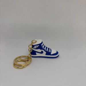 Blå och vit Nike Jordan 1 nyckelring - Nyckelring formad som en Nike Jordan 1 sneaker i blått och vitt med klassisk high-top siluett och detaljerad logga på sidan. Perfekt accessoar för sneakerheads som vill visa sin stil även på nyckelknippan.