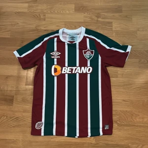 Randig Fluminense fotbollströja Umbro 120 års jubileum  - Säljer en officiell Fluminense fotbollströja från Umbro, jubileumsmodell 120 år. Tröjan har breda vertikala ränder i vinrött, grönt och vitt, med klubbmärke broderat på bröstet och Betano sponsortryck. Rund hals och korta ärmar, tillverkad i lätt polyester.