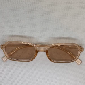 Beige rektangulära solglasögon - Snygga solglasögon med transparent beige båge och rektangulära linser i matchande ton. Glasögonen har en stilren och modern look med tunn plastbåge som ger en lätt känsla. Perfekta för dig som vill ha en trendig accessoar med retrovibbar.