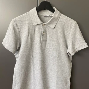 Grå pikétröja från MNG Mango - Snygg grå pikétröja från MNG i storlek XS. Klassisk krage och två knappar framtill. T-shirten har korta ärmar och är gjord i mjuk bomull, perfekt för en chill och stilren look. Passformen är normal och färgen är ljusgrå.