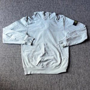Ljusgrå hoodie från Stone Island - Snygg ljusgrå hoodie från Stone Island med klassisk logga på ärmen. Tröjan har huva, magficka och ribbade muddar. Perfekt för dig som gillar streetwear och vill ha något stilrent men ändå med tydlig branding. Sitter som en storlek s. Nypris 3800kr 