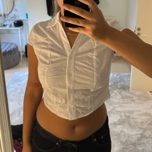Vit croppad skjorta med kort ärm - Säljer en vit croppad skjorta med korta ärmar och klassisk krage. Skjortan har knappar framtill och är figursydd för en snygg passform. Perfekt till jeans eller kjol för en trendig look. Från cider storlek M, liten i storleken. 