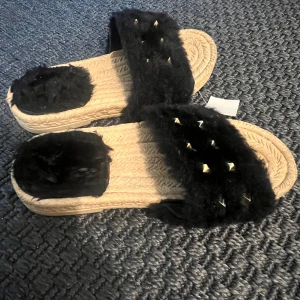 Svarta fluffiga sandaler med nitar - Sandaler med beige flätad sula och bred svart fluffig rem över foten, dekorerad med små silvriga nitar. Snygg och trendig look med mjuk pälsimitation upptill och platt sula. Perfekta för att sticka ut under varma dagar.