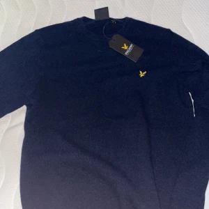 Mörkblå stickad tröja Lyle & Scott - Mörkblå stickad tröja från Lyle & Scott i lammull. Klassisk rund halsringning och gul broderad logga på bröstet. Perfekt för dig som gillar stilren och enkel design med en touch av premiumkänsla.