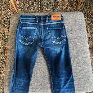 Replay Anbass blå jeans slim fit - Snygga blå jeans från Replay, modell Anbass med slim fit. Jeansen har klassisk femficksdesign, ljusa slitningar och kontrastsömmar. Märkespatch bak i midjan och detaljerade bakfickor. Perfekta för dig som gillar en modern och stilren look.