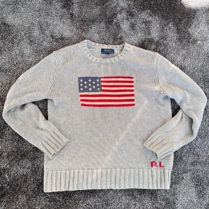 Grå stickad tröja Polo Ralph Lauren - Snygg grå stickad tröja från Polo Ralph Lauren med amerikansk flagga broderad på bröstet och RL-broderi i rött vid nederkanten. Rund hals och långa ärmar med breda ribbade muddar. Perfekt för dig som gillar klassisk stil med en twist.