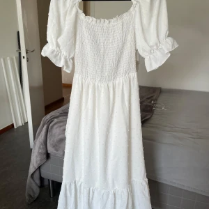 Vit långklänning med puffärm från SHEIN - Superfin vit långklänning från SHEIN i storlek S. Klänningen har puffärmar med volang, smock upptill och ett snyggt knyt i ryggen som ger en öppen rygg. Materialet är 100% polyester med små broderade prickar för extra detalj. Perfekt för sommarens alla tillfällen!