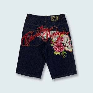 Christian Audigier jeansshorts med broderi - Unika mörkblå jeansshorts från Christian Audigier med fet broderad design på baksidan – döskalle, röda rosor och stora röda bokstäver. Klassisk femficksmodell, gul logga i midjan och detaljerade sömmar. Perfekt för dig som vill sticka ut.