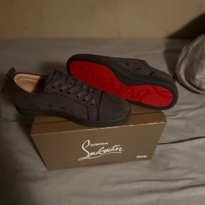 Christian Louboutin skor - Snygga mörkgrå sneakers från Christian Louboutin med klassisk röd sula och rund tå. Skorna är tillverkade i mjuk mocka och har snörning framtill. Perfekt för dig som gillar exklusiva detaljer och vill sticka ut med en ikonisk design.