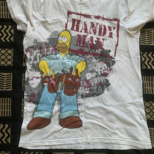 Vit Simpsons t-shirt med Homer-print - Cool vit t-shirt med stor färgglad print av Homer Simpson som 'Handy Man' och text i rött. Trycket har detaljer som verktygsbälte, donuts och Duff-öl. T-shirten har rund hals och korta ärmar, perfekt för dig som gillar humor och popkultur. Men den har ingen etikett 
