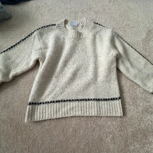Beige stickad tröja från Lindex - Mysig beige stickad tröja från Lindex med svarta detaljer längs ärmar och nederkant. Tröjan har ribbad halsringning och muddar, samt långärmad passform. Tröjan är i 128 men passade på mig som 140-146. Perfekt för kyliga dagar och enkel att matcha med jeans eller kjol.