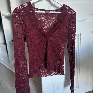 Vinröd spetsblus från Gina Tricot - Säljer en vinröd långärmad spetsblus från Gina Tricot. Blusen har v-ringning, är genombruten med blommigt spetsmönster och har en snygg twistdetalj framtill. Perfekt för dig som vill sticka ut med en romantisk vibe.