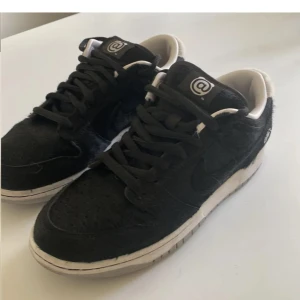 Nike SB Dunk Low svart/vit @logo - Unika Nike SB Dunk Low sneakers i svart och vitt med @-logga på plös och häl. Ovandel i pälsliknande syntetmaterial, vita detaljer och grå sula. Klassisk snörning och låg profil. Perfekta för dig som vill sticka ut med en streetstyle-look.