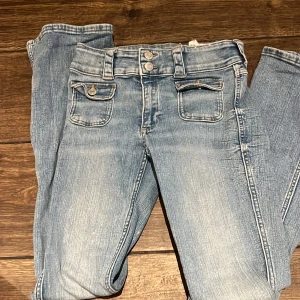 Ljusblå lowasist bootcut jeans med dubbla knappar - Säljer ett par ljusblå bootcut jeans med dubbla knappar i midjan och dekorativa fickor fram. 