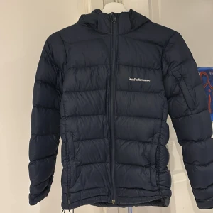 Mörkblå dunjacka från Peak Performance - Säljer en mörkblå dunjacka från Peak Performance med huva och dragkedja framtill. Jackan har två sidofickor med dragkedja och är perfekt för kyligare dagar. Klassisk och stilren design med logga på bröstet. Använd fåtals gånger säljer för att den är alldeles för liten. Köptes för 2900kr på skitstarshop hemsida. Storlek XS/160. Pris kan diskuteras!