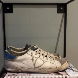 Vita sneakers från Philippe Model med blå detalj - Säljer ett par vita sneakers från Philippe Model med blå detalj på hälen och gråa partier. Skorna har snörning och en platt sula. Perfekta för dig som gillar klassisk och sportig stil.