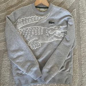 Grå sweatshirt med krokodiltryck från Lacoste - Säljer en grå sweatshirt från Lacoste med stort vitt krokodiltryck över bröstet. Tröjan har rund halsringning och ribbade muddar vid ärmslut och nederkant. Perfekt för en avslappnad och sportig stil.