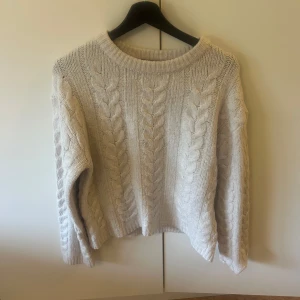 Beige stickad tröja från 157 - Mysig beige stickad tröja från 157 med flätstickat mönster och rund halsringning. Tröjan har långa ärmar och ribbade muddar vid hals, ärmslut och nederkant. Perfekt för kyliga dagar. Storlek S