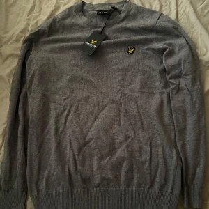 Grå tröja från Lyle & Scott - Säljer en klassisk grå tröja från Lyle & Scott med rund halsringning och deras ikoniska gula logga broderad på bröstet. Tröjan har långa ärmar och ribbade muddar vid ärmslut och nederkant. Perfekt för en stilren look.
