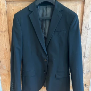 Svart kostym från Dressmann Slim Fit - Säljer en svart kostym från Dressmann i slim fit-modell. Kavajen har klassisk krage, två knappar framtill och fickor med lock. Byxorna har pressveck och normal midja. Perfekt för bal eller fest.