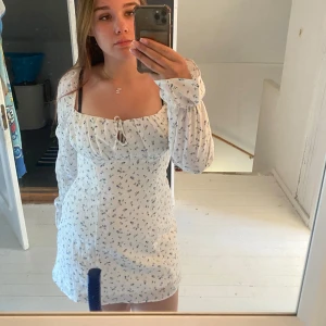 Vit blommig kortklänning med lång ärm - Söt vit kortklänning med små blå blommor och långa ärmar. Klänningen har knyt fram och volangdetaljer vid ärmslut. Den har en feminin passform och öppen rygg. Perfekt för skolavslutningen eller sommarfesten! Pris kan diskuteras, endast använd en gång!