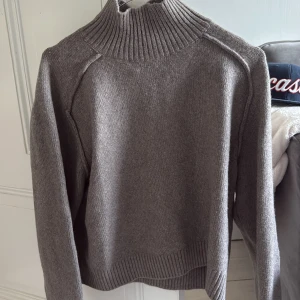 Grå stickad polotröja - Säljer en grå stickad polotröja med hög krage och långa ärmar. Tröjan har en ribbad struktur vid hals, ärmslut och nederkant. Perfekt för kyligare dagar och enkel att matcha med olika outfits.