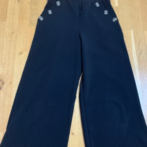 Svarta vida halvshorts  med knappar från Primark - Svarta halvshorts från Primark med breda ben och dekorativa knappar framtill vid midjan. Shortsen har hög midja och en klassisk, stilren look som passar till många olika outfits.