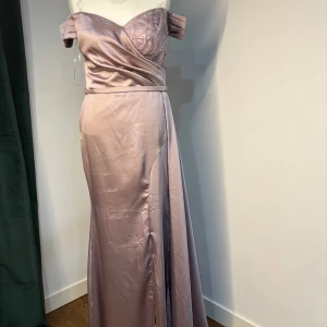 Ljuslila långklänning i satin med offshoulder - Ljuslila långklänning i glansigt satinmaterial med offshoulder-ärmar och omlottliv. Klänningen har en hög slits framtill och vacker drapering över bysten. Perfekt för bal eller fest. Dragkedja i ryggen för enkel påklädning.