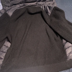 Svart dunväst från Moncler med stickade detaljer - Säljer en svart dunväst från Moncler med stickade ärmar och sidor. Västen har dragkedja framtill, två fickor med knappar och Moncler-logga på ärmen. Perfekt för lager-på-lager och snygg till många outfits. Storlek M men passar i L för den e lång vid armarna 