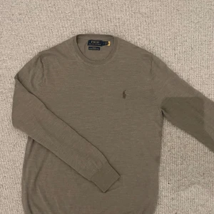 Beige marinoulltröja från Polo Ralph Lauren - Säljer en stilren beige tröja från Polo Ralph Lauren i 100% merinoull. Klassisk rund halsringning och diskret broderad logga på bröstet. Perfekt för en snygg och enkel look.