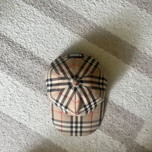 Beige rutig keps från Burberry - Säljer en klassisk keps från Burberry i beige med det ikoniska rutiga mönstret i svart, vitt och rött. Kepsen har böjd skärm och justerbar rem baktill. Perfekt accessoar för en stilren look.