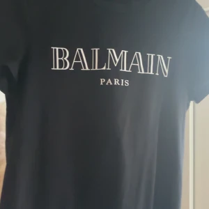 Svart t-shirt från Balmain Paris - Snygg svart t-shirt från Balmain Paris med vit logga tryckt på bröstet. Klassisk passform och rund halsringning. Perfekt för dig som gillar stilrena märkesplagg.