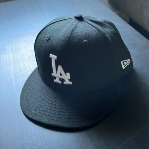 Svart LA-keps från New Era - Säljer en svart keps från New Era med LA-logga framtill i vitt. Klassisk modell med böjd skärm och broderad logotyp på sidan. Kepsen är i storlek 7 1/8 (56,8 cm) och passar perfekt för dig som gillar att en skön stil. Pris kan diskuteras vid en snabb affär 