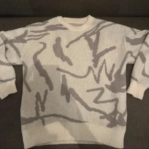 Beige och grå stickad tröja med abstrakt mönster - Säljer en beige stickad tröja med grått abstrakt mönster. Tröjan har rund halsringning, långa ärmar och ribbade muddar vid ärmslut och nederkant. Perfekt för dig som gillar unika plagg med cool design.