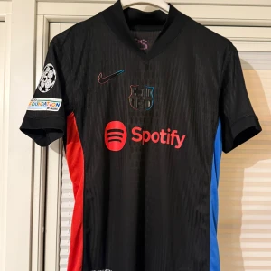 FC BARCELONA TRÖJA LAMINE YAMAL 19 BORTASTÄLL 2024/2025 - Säljer en svart FC Barcelona bortatröja med röda och blå detaljer, sponsorlogga från Spotify och Champions League-märke på ärmen. Tröjan har tryck med Lamine Yamal och nummer 19 på ryggen. Klassisk Nike Dri-FIT-modell. Kör ej via Plick utan kör utanför appen. Swish och kan även fraktas! Storlek: M.