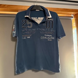 Blå pikétröja från Camp David - Säljer en blå pikétröja från Camp David med tryck och broderade detaljer fram och bak. Tröjan har korta ärmar, vit och blå krage samt knappar vid halsen. Perfekt för en sportig och avslappnad stil.