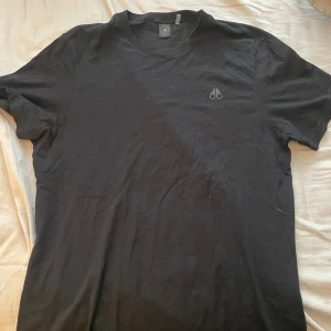 Svart t-shirt från Moose Knuckles - Säljer en svart t-shirt från Moose Knuckles med diskret logga på bröstet och tryckt varumärkesnamn på ärmen. Klassisk passform och rund halsringning. Perfekt för en stilren och enkel look.oanvänd köpt för 1500