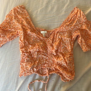 Söt topp  - Säljer en blommig croppad blus i orange och vit med v-ringning och knyt framtill. Blusen har trekvartsärm och ett rynkat parti i mitten som ger en snygg passform. Perfekt för vår och sommar!
