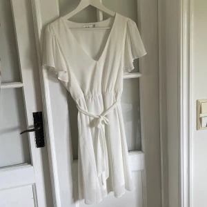 Vit omlottklänning från VILA - Säljer en vit kortklänning från VILA i storlek 36. Klänningen har v-ringning, korta volangärmar och knytskärp i midjan. Perfekt för student/skolavslutning.