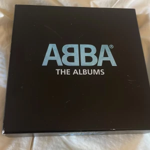 ABBA The Albums box - Säljer en svart box med texten 'ABBA The Albums' i blått och vitt. Boxen innehåller flera album av ABBA, bland annat 'Arrival' med ikoniskt omslag. Perfekt för dig som älskar musik och vill ha en samling med klassiska ABBA-album.❤️
