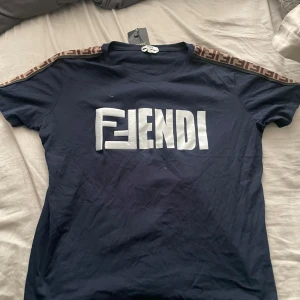 Mörkblå t-shirt med logga från Fendi - Säljer en mörkblå t-shirt från Fendi med stor vit logga på bröstet och bruna detaljer med Fendi-mönster längs axlarna. Klassisk rund hals och korta ärmar. Perfekt för dig som gillar streetwear och designerplagg. Helt ny köpt för 3500 